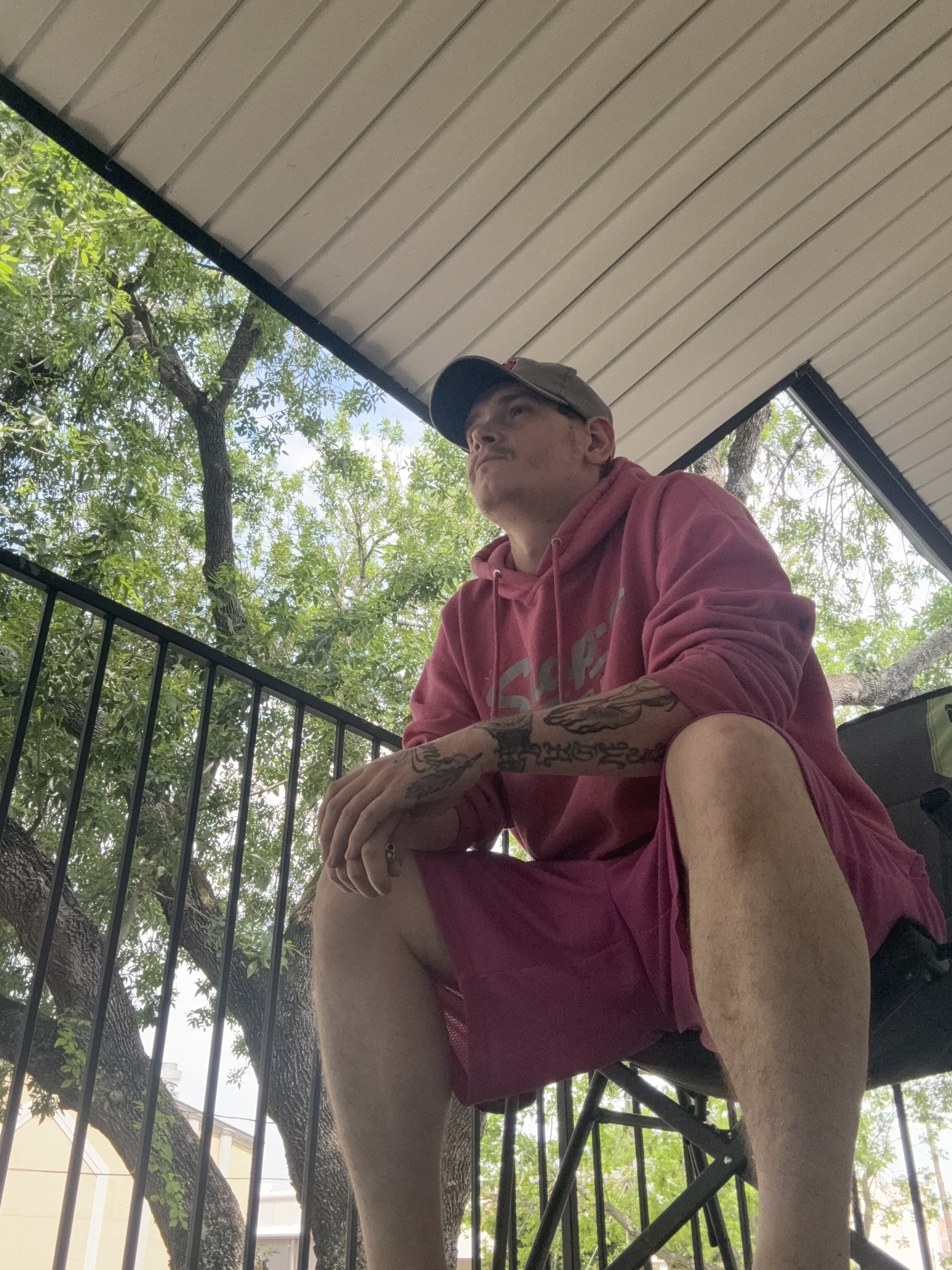 KoCain in a pink hoodie and gray cap on an outdoor balcony, tattooed arms folded in front