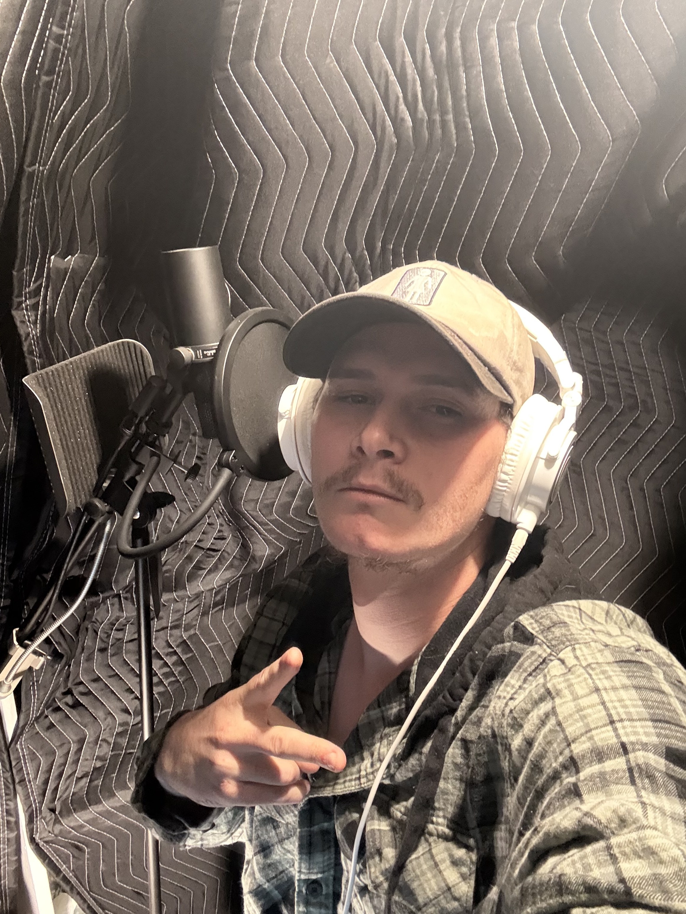 KoCain in a recording booth, gray cap and white headphones, throwing a hand sign at the camera