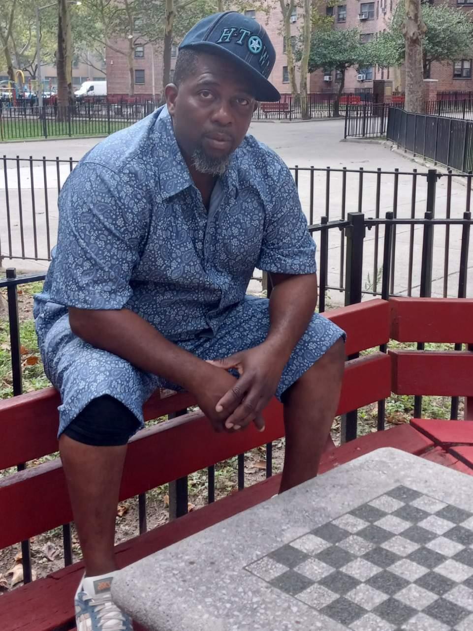 Mr. Shorts seated on a red park bench in a blue floral set and HTOWN cap, near a chessboard table