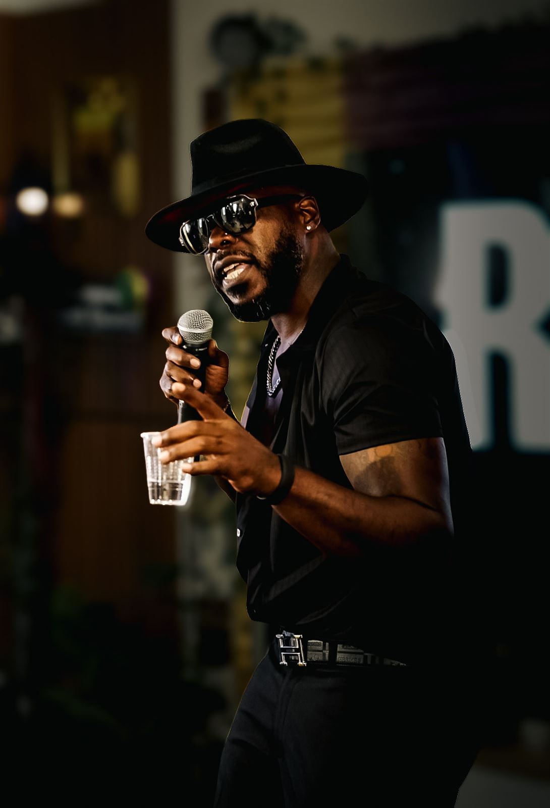 Teknition at Rhythm & Rise Live 2026, in black fedora and sunglasses singing into the mic while holding a cup