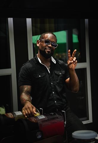 Behind the scenes at RRL 2026, bartender in black shirt and sunglasses flashing a peace sign behind the drink station