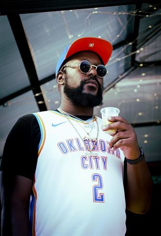 Big Tree #4real at RRL 2026, artist in orange-brim cap, round sunglasses and Oklahoma City jersey holding drink under string lights