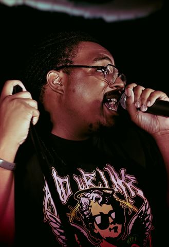 Einstein the Mastermind at RRL 2026, in glasses and black cherub graphic tee singing into mic under warm red-tinted stage light
