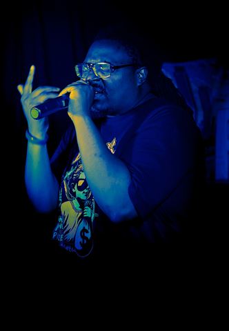 Einstein the Mastermind at RRL 2026, in glasses rapping into handheld mic under deep blue stage light with hand raised