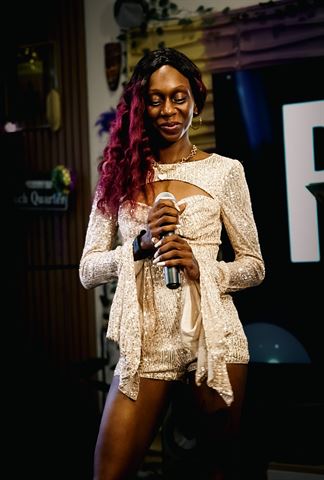 Ife Devine at RRL 2026, long burgundy hair and sparkly sequin romper, holding the mic in warm indoor lighting