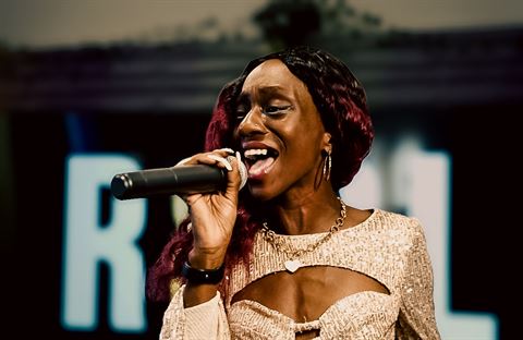 Ife Devine at RRL 2026, close-up with burgundy hair and gold necklace singing into the mic before glowing letters