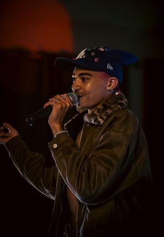KaliiFreak at RRL 2026, in jeweled blue cap and fur-collared jacket smiling into the mic with warm glow behind