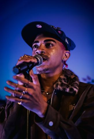 KaliiFreak at RRL 2026, in jeweled cap with rings and layered necklaces singing into the mic on the deep blue stage