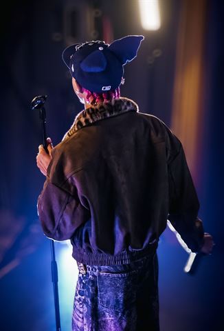 KaliiFreak at RRL 2026, from behind in eared cap gripping the mic stand in backlit haze