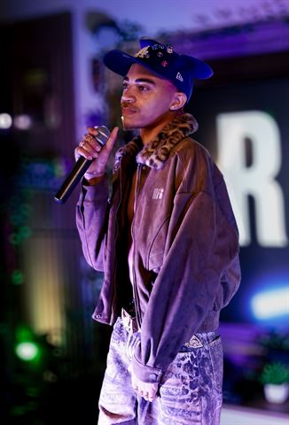 KaliiFreak at RRL 2026, in jeweled blue cap and fur-collared jacket holding the mic beside the letter sign