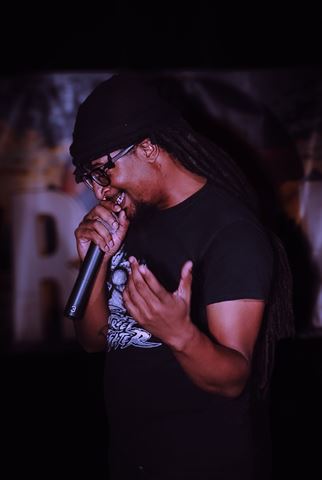 MayB.L8ter at RRL 2026, man with locs in black durag and glasses rapping into handheld mic under warm stage light