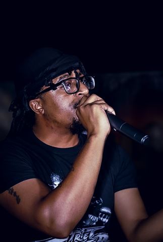 MayB.L8ter at RRL 2026, close-up in glasses and black durag gripping mic with both hands in dim lighting