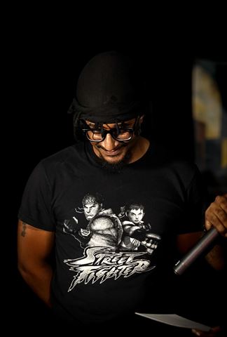 MayB.L8ter at RRL 2026, smiling in Street Fighter tee and black durag looking down with mic in hand