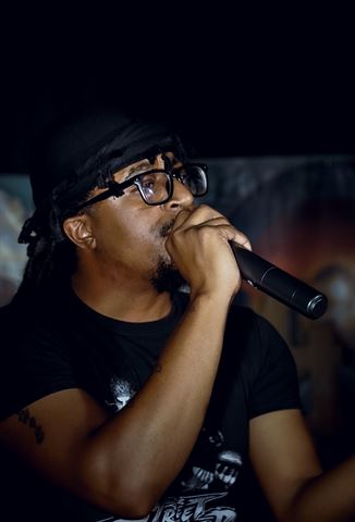 MayB.L8ter at RRL 2026, close-up in glasses and black durag singing into mic against blurred dark backdrop