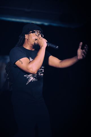 MayB.L8ter at RRL 2026, man in black graphic tee and glasses rapping into mic with arm outstretched on dark stage