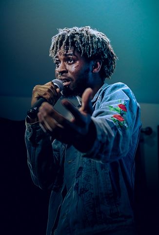 Mell. at RRL 2026, in rose-embroidered denim jacket rapping into the mic, reaching toward the camera in teal light