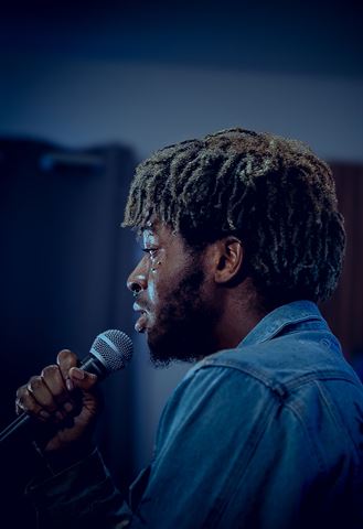 Mell. at RRL 2026, profile with dreads in denim jacket singing into handheld mic under blue light