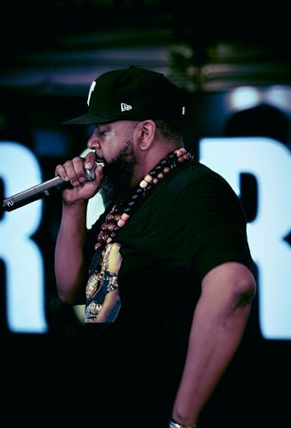 Mirage the Gr8 at RRL 2026, bearded artist in black ball cap and wooden bead necklaces rapping before lit letter signage