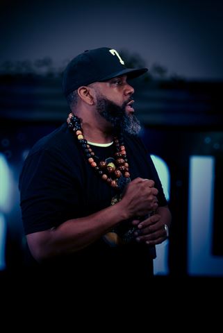 Mirage the Gr8 at RRL 2026, in black cap and layered bead necklaces clasping mic under cool blue light