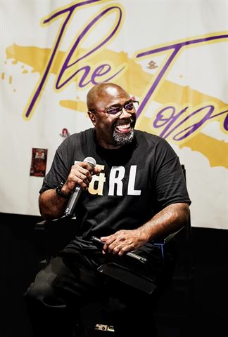 Mr Durite, laughing host in glasses and black R&RL shirt holding microphone before painted banner