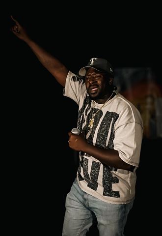PushPlay914 at RRL 2026, in cap and graphic tee with arm raised high, holding the mic on the dark stage