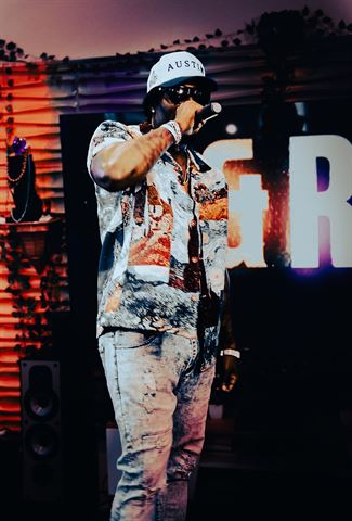 Tay No Tee at RRL 2026, in white Austin cap and patterned shirt rapping onstage before the lit letter backdrop