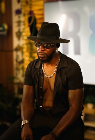 Teknition at RRL 2026, seated in black fedora and open shirt with chunky silver chain in warm window light