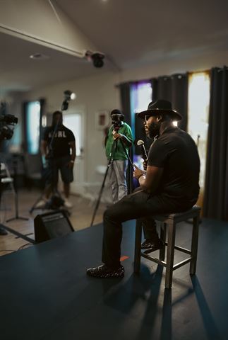 Teknition at RRL 2026, in fedora seated on a stool with mic, facing the camera crew in the bright room