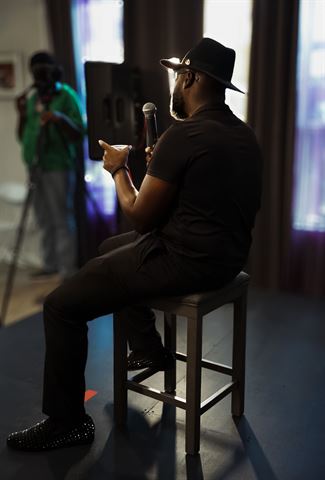 Teknition at RRL 2026, in fedora seated on a stool holding mic and cup, backlit by bright windows