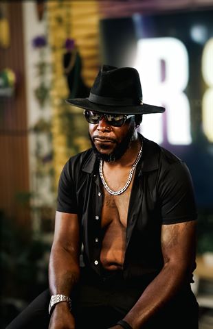 Teknition at RRL 2026, in black fedora, sunglasses and open black shirt with silver chain, seated