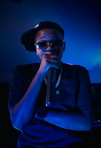 TGN Lank$ at RRL 2026, in tilted cap, sunglasses and chain holding the mic close under deep blue light