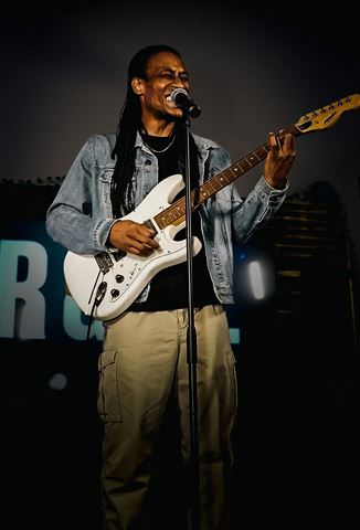Yadin Kol at RRL 2026, guitarist with locs in denim jacket singing before the lit R&RL sign
