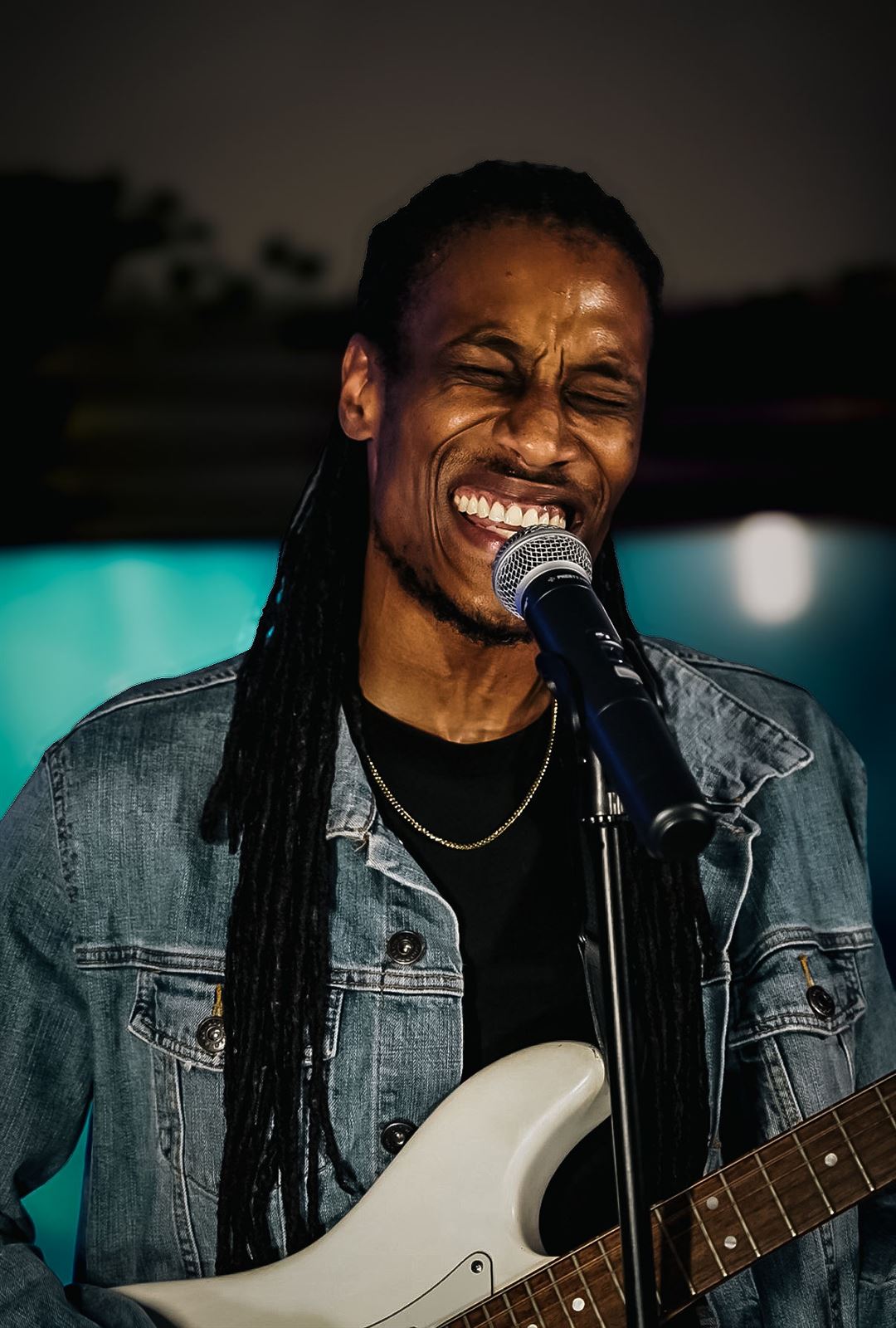 Yadin Kol at Rhythm & Rise Live 2026, with long locs in denim jacket smiling at the mic with white guitar in teal glow
